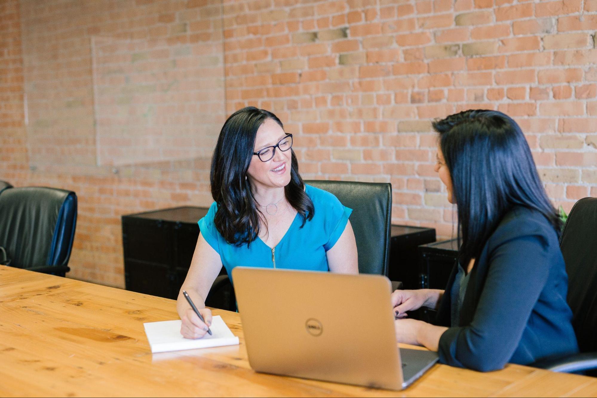 Two women are sitting at a wooden table in a meeting room with a brick wall background. One woman, likely discussing blockchain technology, is holding a pen and notepad, smiling, and facing a laptop. The other woman is engaging with her, also smiling. Both are wearing business attire.