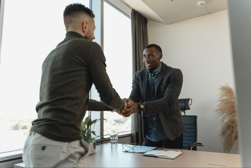 Two men are shaking hands across an office desk. One is standing with a smile, wearing a blazer and jeans. The other, seen from the back in a sweater and light-colored pants, is finalizing their agreement. With large windows lighting up the room's potted plant and open notebook, they're discussing effective strategies for recruiting top talent.